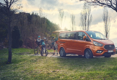 Drei Kinder laden ihre Fahrräder in den Kofferraum eines orangenen Ford Tourneo Custom