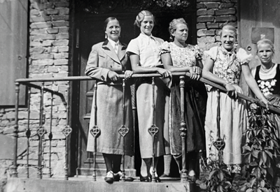 Fünf Frauen in Kleidern stehen aufgereiht auf einer Treppe vor einem Haus