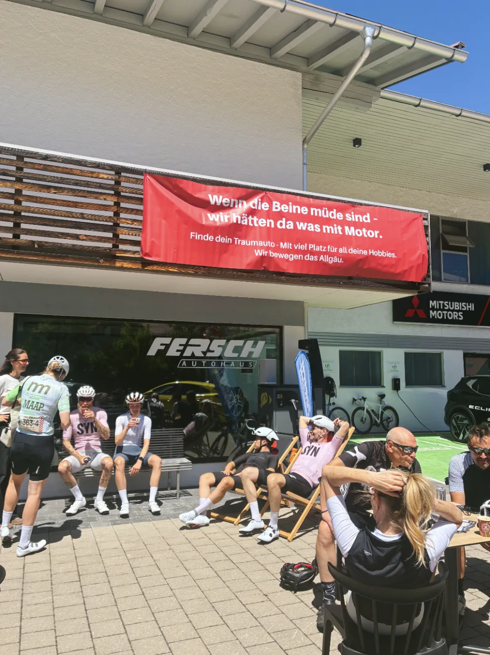 Radfahrer machen Pause vor dem Autohaus Fersch, darüber das Banner: Wenn die Beine müde sind, wir hätten da was mit Motor